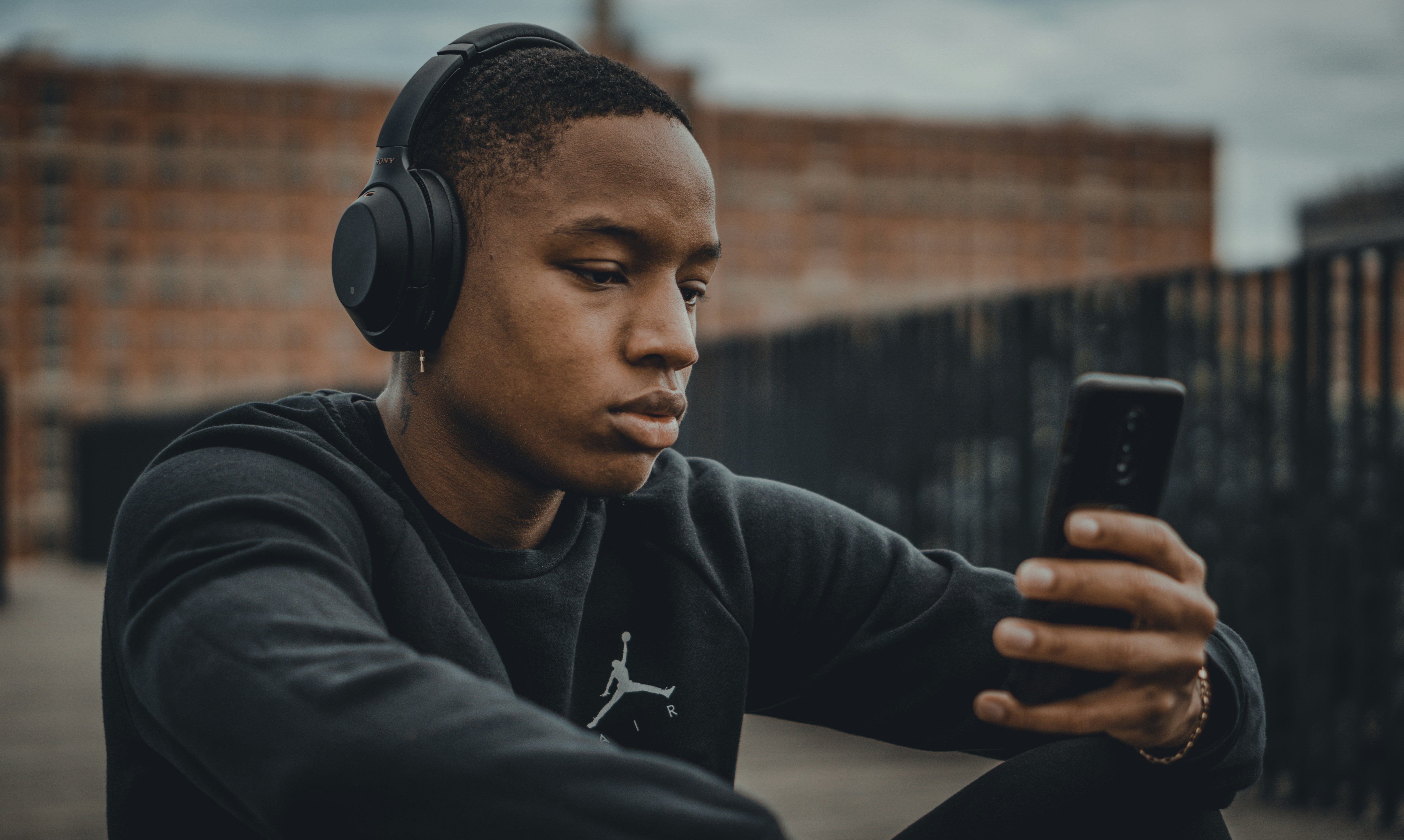 Man in a black nike hoodie sitting down wearing black headphones & holding a mobile phone.